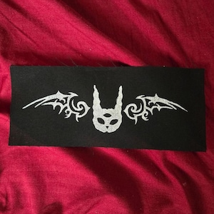 May include: A rectangular black fabric patch featuring a centered silver-colored graphic print. The design depicts a stylized creature head with three eyes and pointed ears, flanked by two symmetrical, intricate wing-like shapes on each side.