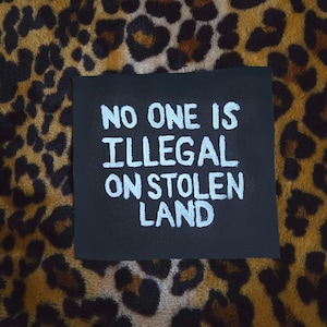 以下が含まれることがあります： 黒い正方形の布製パッチで、白い刺繍で「NO ONE IS ILLEGAL ON STOLEN LAND」というテキストが施されています。文字は太字のサンセリフ体で、暗い背景の中央に配置されており、高いコントラストを生み出しています。