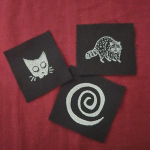 May include: Three square black fabric patches featuring white embroidered designs. One patch displays a stylized cat head, another shows a detailed raccoon, and the third features a large, centered spiral pattern. Each patch has a clean edge.