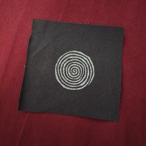 May include: A square black fabric patch featuring a white embroidered spiral design centered on the material. The patch has a visible woven texture and clean, straight edges, showcasing a simple circular pattern stitched in white thread.
