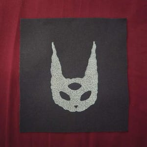 May include: A square black fabric patch featuring a centered, light gray, textured graphic of a three-eyed creature head with tall, pointed ears. The design is screen-printed onto the dark textile, creating a high-contrast visual element.