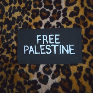 Puede incluir: Un parche rectangular de tela negra con el texto bordado en blanco FREE PALESTINE en una fuente sans-serif. El parche está colocado sobre un fondo con estampado de leopardo en tonos marrón, negro y beige con una textura suave y peluda.
