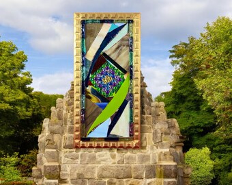 Könnte beinhalten: Ein farbenfrohes Buntglasmosaik-Kunstwerk in einem goldenen Rahmen, das in eine Steinmauer eingelassen ist. Das Mosaik zeigt geometrische Formen in Blautönen, Grün- und Brauntönen, mit einem zentralen kreisförmigen Design aus schillernden Glasperlen.