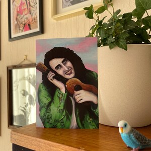 Pode incluir: Uma impressão de arte colorida de uma pessoa com cabelo longo e escuro, usando uma jaqueta verde e segurando um ukulele. O fundo é rosa e azul. Uma pequena figura de pássaro azul e branco está em uma superfície de madeira.