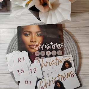 May include: A Christmas Bingo game set featuring a woman's portrait and the word "Christmas" in gold. Includes bingo cards, number cards, and prize cards. The set is arranged on a gray woven mat.