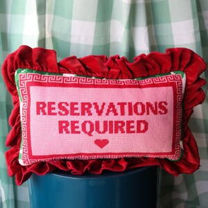 Pu&ograve; includere: Un cuscino decorativo con un centro rosa con la scritta "RESERVATIONS REQUIRED" in rosso, incorniciata da un bordo rosso e verde. Il cuscino &egrave; circondato da un bordo di velluto rosso arricciato.