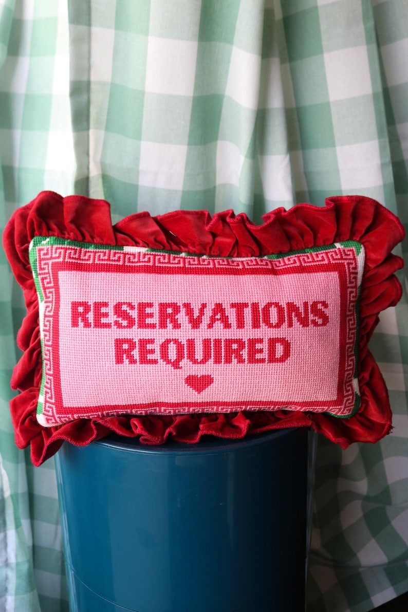 Pu&ograve; includere: Un cuscino decorativo con un centro rosa con la scritta "RESERVATIONS REQUIRED" in rosso, incorniciata da un bordo rosso e verde. Il cuscino &egrave; circondato da un bordo di velluto rosso arricciato.