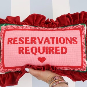 Pu&ograve; includere: Un cuscino rettangolare rosa ricamato con le parole "RESERVATIONS REQUIRED" in rosso. Presenta un cuore rosso e un bordo rosso e verde. Il cuscino &egrave; circondato da un bordo di velluto rosso arruffato.