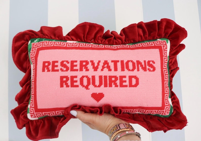 Pu&ograve; includere: Un cuscino rettangolare rosa ricamato con le parole "RESERVATIONS REQUIRED" in rosso. Presenta un cuore rosso e un bordo rosso e verde. Il cuscino &egrave; circondato da un bordo di velluto rosso arruffato.