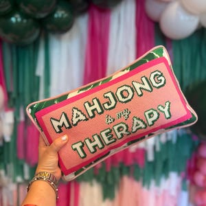 May include: A rectangular pink pillow with the text "MAHJONG is my THERAPY" in white and green. The pillow has a green border and a tropical leaf pattern. The pillow is being held by a person.