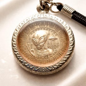 May include: A silver pendant with an alien design, encased in a clear, round setting. The alien figure has large eyes and is displaying a peace sign. The pendant is attached to a black cord.