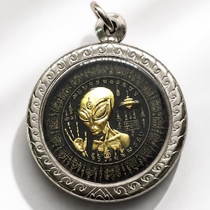 Puede incluir: Un colgante circular de tono plateado que presenta una figura alienígena dorada con una mano levantada y un pequeño platillo volante. El fondo es negro con intrincadas escrituras y símbolos dorados, enmarcado en metal plateado.