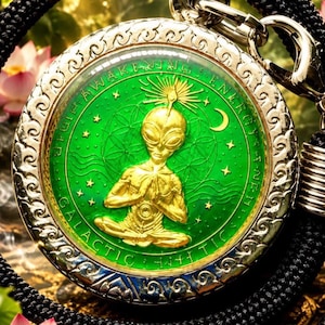 Può includere: Un ciondolo circolare raffigurante una figura aliena color oro in posa meditativa su uno sfondo verde brillante. Il bordo è inciso con il testo AWAKENING ENERGY e GALACTIC, arricchito da stelle e una mezzaluna. È appeso a un cordino nero.