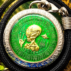 Puede incluir: Un colgante circular que presenta una figura alienígena dorada y un platillo volante sobre un fondo verde vibrante con intrincados símbolos grabados. El colgante está encerrado en un marco metálico con detalles azules y cuelga de un cordón negro.