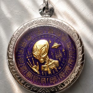 Può includere: Un ciondolo circolare color argento con una figura aliena dorata al centro con una mano alzata e un piccolo disco volante. Lo sfondo è una superficie viola intenso coperta da intricate scritte e simboli geometrici dorati.