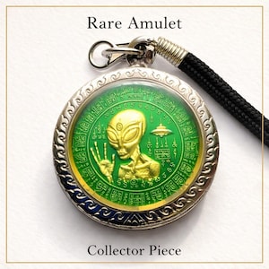 Op de afbeelding: Een zilverkleurige amulet met een groen en goudkleurig alien-ontwerp. De amulet heeft een gedetailleerde rand en een zwart koord. Tekst op de amulet luidt "Rare Amulet" en "Collector Piece".