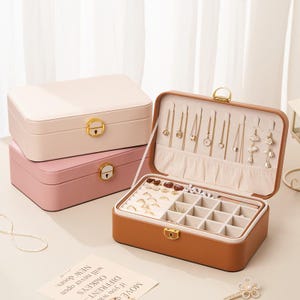 Peut inclure: Trois boîtes à bijoux de couleurs rose et beige. La boîte supérieure est rose clair, celle du milieu est rose foncé et la boîte inférieure est beige. La boîte beige est ouverte, révélant des compartiments pour bijoux. Fermoirs et ferrures dorés visibles.