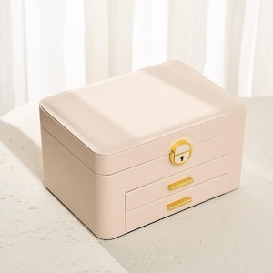 May include: A rectangular jewellery box in a soft cream colour with a smooth finish. The box features a gold-toned metal latch with a keyhole on the top lid and two horizontal drawers below, each equipped with a slim, matching gold-toned metal handle.