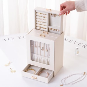 May include: A cream-coloured jewellery box with multiple compartments and drawers. The top section has ring slots and earring holders. The middle section has a glass door to display necklaces. The bottom drawer is open, revealing two bracelets. Gold-coloured hardware accents the box.