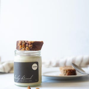 May include: A glass jar with a black label that reads "daily bread" with the ingredients listed below. A slice of banana bread topped with walnuts sits on top of the jar. A second slice of bread is on a plate.