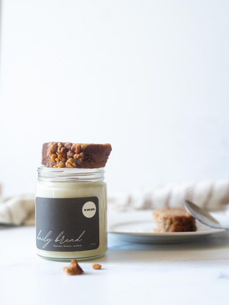 May include: A glass jar with a black label that reads "daily bread" with the ingredients listed below. A slice of banana bread topped with walnuts sits on top of the jar. A second slice of bread is on a plate.