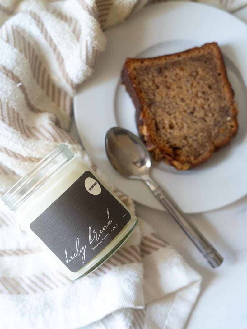 May include: A glass jar candle with a black label that reads "daily bread" in white script, next to a slice of banana bread on a white plate with a spoon. The candle is on a striped beige and white towel.