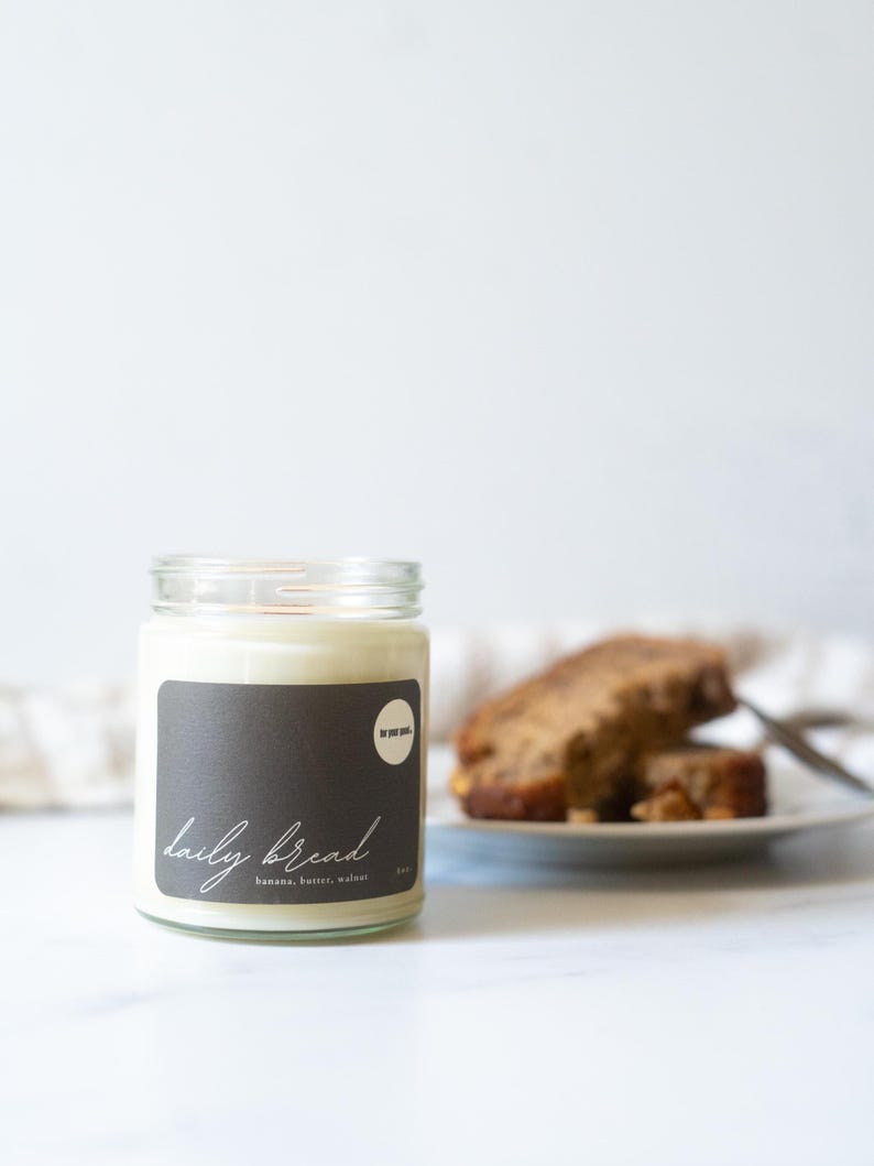 May include: A glass jar candle with a dark gray label that reads "daily bread" in cursive. The label also lists "banana, butter, walnut." Slices of banana bread sit on a white plate in the background.