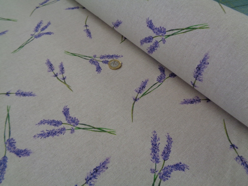May include: A light beige fabric featuring a repeating pattern of lavender sprigs in purple and green. The fabric is folded, revealing its texture and design. A coin is visible on the fabric.