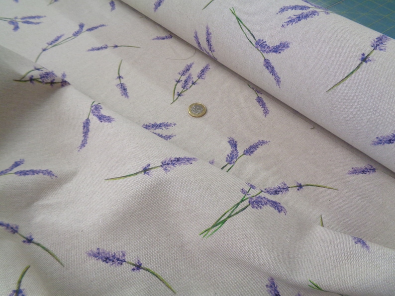 May include: A close-up of fabric featuring a lavender sprig pattern. The lavender is purple with green stems, set against a light beige backdrop. The fabric is folded, revealing its texture. Ideal for crafting or home decor projects.