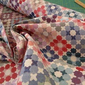 May include: A close-up of a fabric with a geometric pattern of multi-coloured flowers. The fabric is woven with a variety of colours, including red, pink, blue, and green. The fabric is folded in half, and the pattern is visible on both sides.