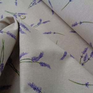 May include: Beige fabric featuring a lavender sprig pattern. The lavender flowers are purple, with green stems. The fabric is folded, revealing its texture and the details of the print. A coin is visible on the fabric.