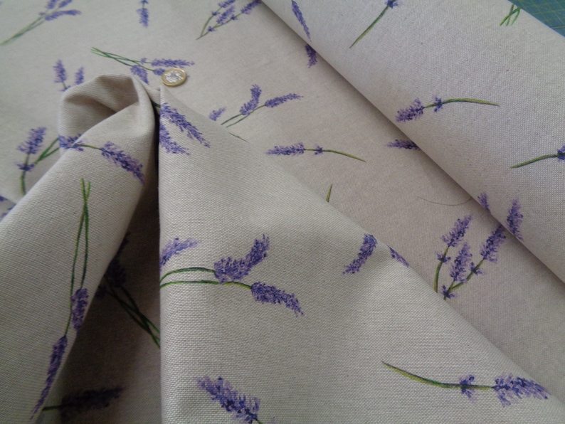 May include: Beige fabric featuring a lavender sprig pattern. The lavender flowers are purple, with green stems. The fabric is folded, revealing its texture and the details of the print. A coin is visible on the fabric.