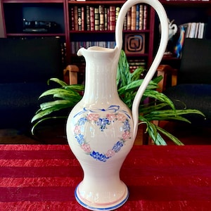 May include: A white ceramic pitcher with a tall, curved handle. The pitcher features a heart-shaped floral design in pink and blue, with a blue bow at the top. The base and rim have pink and blue accents.