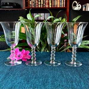 May include: Four clear glass goblets with a beaded stem and flared rim are arranged on a blue surface. Pink flowers are in the foreground. The goblets are in front of green plants and a bookshelf.