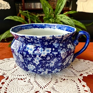 May include: A ceramic chamber pot featuring a deep blue and white floral pattern. The vessel has a wide, scalloped rim, a rounded body, and a single curved handle on the side. The interior is white, and the exterior is covered in a dense, intricate botanical design of small blossoms and foliage.
