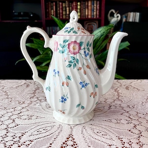 May include: White ceramic teapot with a lid, featuring a floral pattern of pink, blue, and green flowers and berries. The teapot has a curved handle and spout, and a decorative swirl design. A classic piece for serving tea.