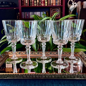 May include: Five clear crystal wine glasses with a ribbed design, displayed on a decorative gold-trimmed tray. The glasses have a classic shape with a long stem and a wide base. The background includes a bookshelf and greenery.