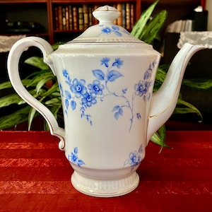 May include: A white porcelain teapot featuring a delicate blue floral pattern with vines and leaves. The teapot has a matching lid with a knob, a curved handle, and a spout, all accented with thin gold trim along the edges and base.