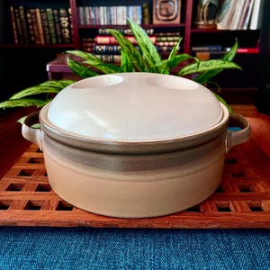 May include: A ceramic casserole dish with a lid. The dish has a light beige and brown gradient design. The lid has two recessed circular areas. The dish has two handles and is sitting on a wooden tray.