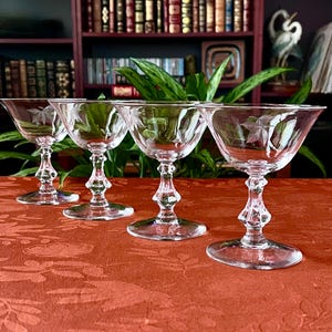 May include: Four clear crystal champagne coupe glasses with etched floral designs. The glasses have a wide bowl, a decorative stem, and a round base. They are arranged on a red tablecloth.