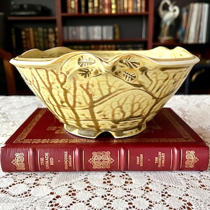 May include: A yellow ceramic bowl with a gold rim and a tree branch design. The bowl is sitting on top of a red book with gold lettering that reads "The Unmade Bed - Francoise Sagan - First Edition - The Franklin Library".
