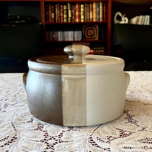 May include: A round ceramic casserole dish with a lid, featuring a two-tone design. The left side is a deep dark brown, while the right side is a light cream color. The lid has a central knob and follows the same split color pattern.