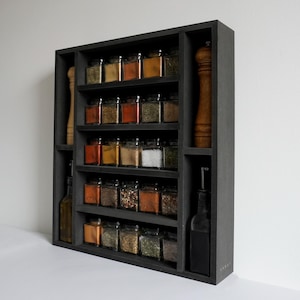 Może przedstawiać: Czarna p&oacute;łka na przyprawy z wieloma p&oacute;łkami wypełnionymi szklanymi słoikami z przyprawami. P&oacute;łka ma dwa pionowe przedziały po bokach, jeden z czarną butelką, a drugi z brązowym młynkiem do pieprzu.