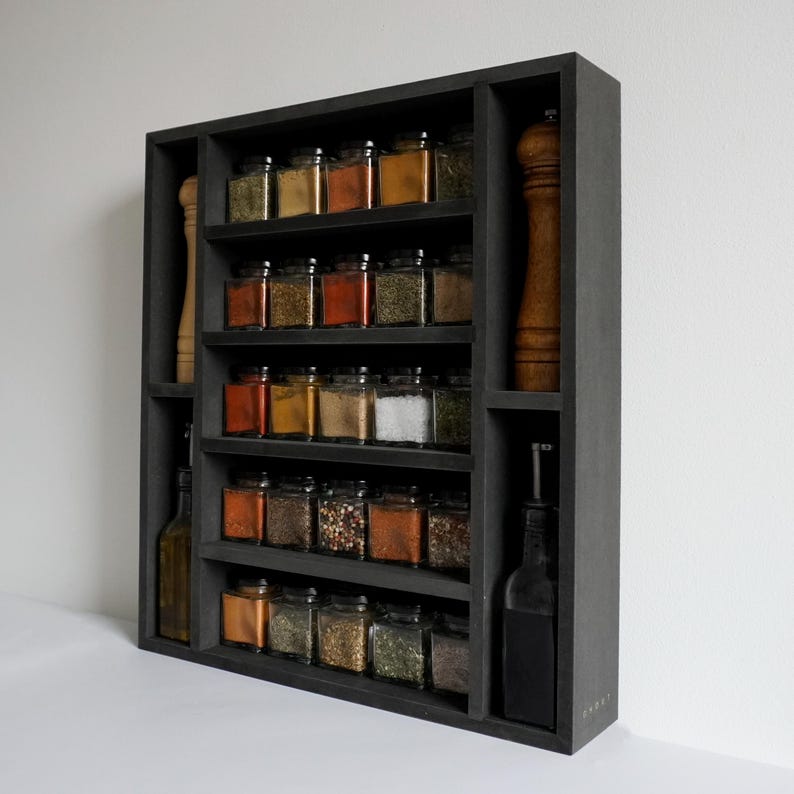 Może przedstawiać: Czarna p&oacute;łka na przyprawy z wieloma p&oacute;łkami wypełnionymi szklanymi słoikami z przyprawami. P&oacute;łka ma dwa pionowe przedziały po bokach, jeden z czarną butelką, a drugi z brązowym młynkiem do pieprzu.