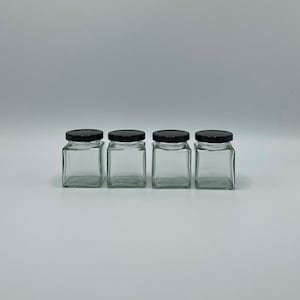 Op de afbeelding: Vier glazen potten met zwarte deksels. De potten zijn vierkant en hebben een gladde, transparante glazen afwerking.