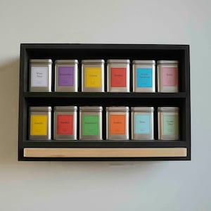 Könnte beinhalten: Ein schwarzes Holzregal mit zwei Ablagen beherbergt zwölf silberne Dosen mit bunten Etiketten. Die Etiketten tragen Text in Niederländisch, darunter "Witte Thee", "Rooibos", "Goede Nachtrust", "Kamille", "Aardbei", "Brandnetel", "Gember", "Mint", "Groene Thee", "Citroen", "Relax" und "Boerenvruchten".