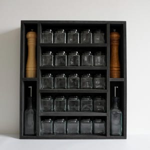 Może przedstawiać: Czarna drewniana p&oacute;łka na przyprawy z pięcioma p&oacute;łkami, na kt&oacute;rych znajduje się 20 przezroczystych szklanych słoik&oacute;w z czarnymi pokrywkami. Po bokach znajdują się dwa drewniane młynki do pieprzu i dwie szklane butelki z olejem.