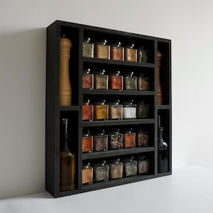 Może przedstawiać: Czarna drewniana p&oacute;łka na przyprawy z wieloma p&oacute;łkami wypełnionymi szklanymi słoikami z przyprawami. P&oacute;łka ma dwa pionowe przedziały po bokach, jeden z drewnianym młynkiem do pieprzu, a drugi ze szklaną butelką oleju.