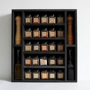 Może przedstawiać: Czarna drewniana p&oacute;łka na przyprawy z pięcioma p&oacute;łkami, każda z małymi szklanymi słoikami z etykietami. Słoiki są wypełnione r&oacute;żnymi przyprawami, w tym papryką, oregano i cynamonem. P&oacute;łka ma r&oacute;wnież dwa otwory na większe butelki, po jednej z każdej strony.