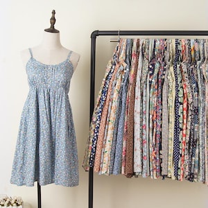 May include: A light blue floral print dress with spaghetti straps on a mannequin. A rack of various patterned fabrics hangs to the right. The dress has a gathered bodice and a flared skirt. The fabrics on the rack feature floral, polka dot, and geometric designs.